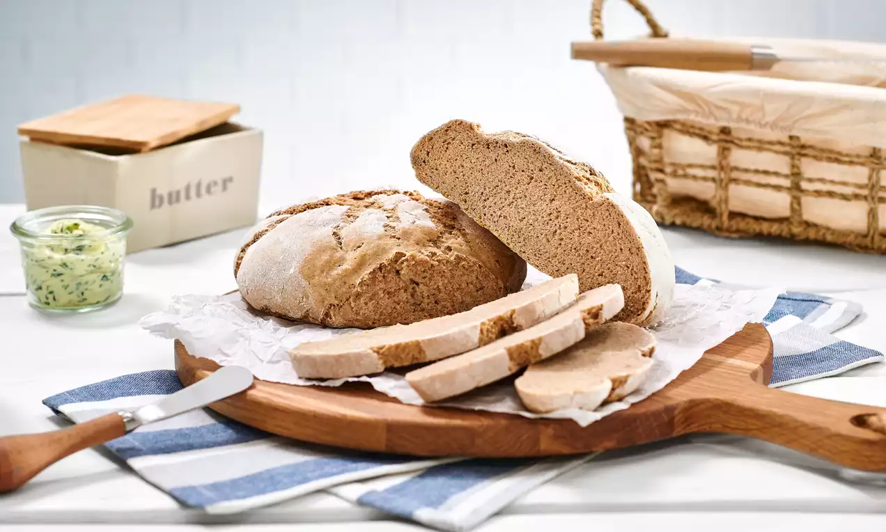 Rundes, goldbraunes Sauerteigbrot mit knuspriger Kruste, halbiert und in Scheiben geschnitten auf einem Holzbrett mit blau-weißem Tuch