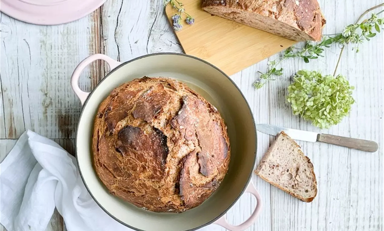 Rundes, goldbraun gebackenes Brot mit rissiger Kruste in einem cremefarbenen Topf, umgeben von Brotstücken, Holzbrett und grüner Deko