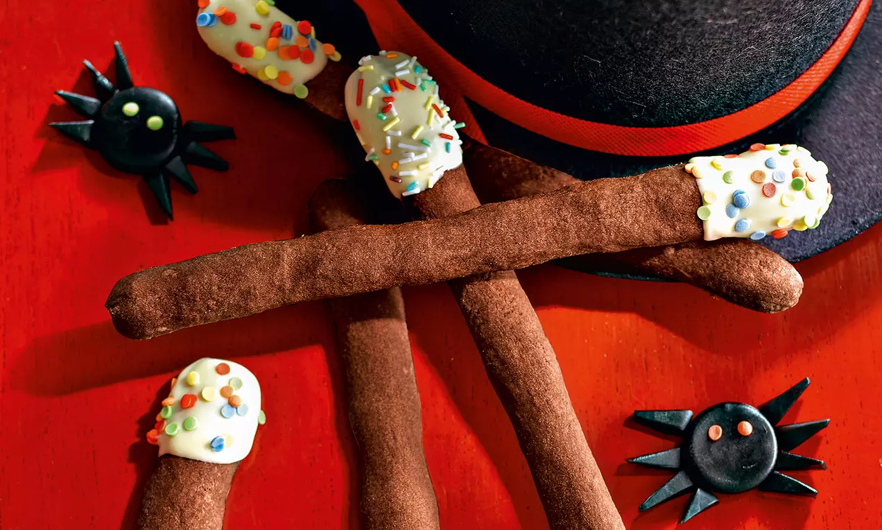 Bastoncini sottili di biscotto al cacao con estremità immerse nel cioccolato bianco, decorati con confettini colorati su tavola rosso acceso accanto a piccoli ragni di caramella