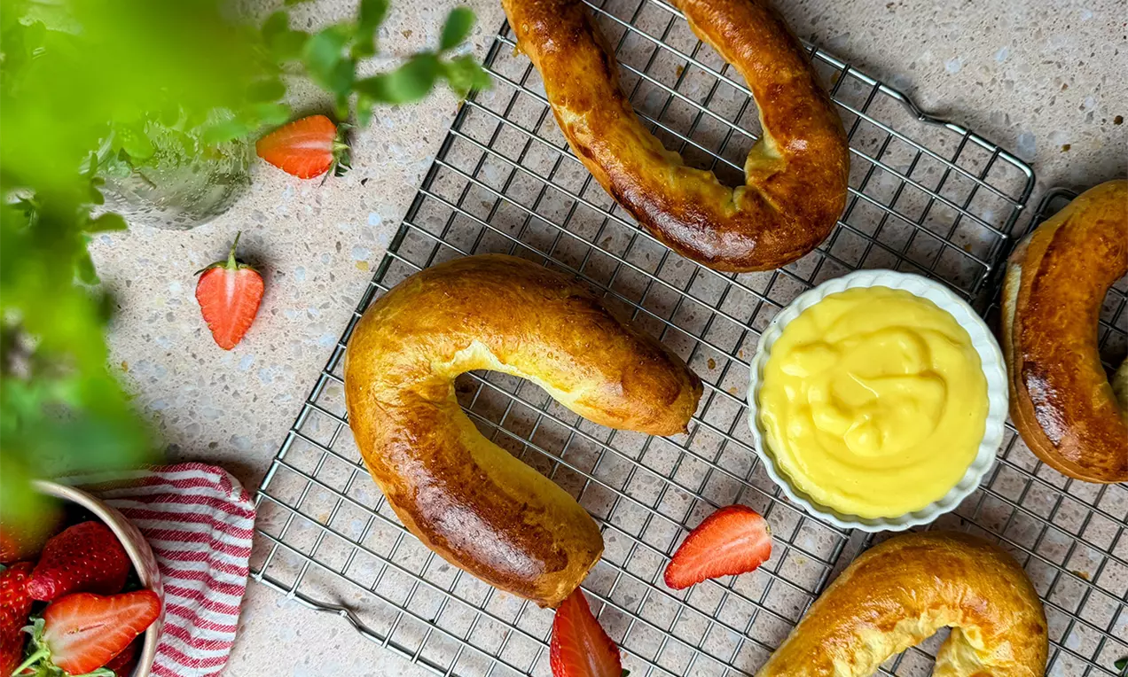 Goldbraune, gebogene Hörnchen auf einem Metallgitter mit gelber Creme in einer weißen Schale und frischen Erdbeerstücken dekoriert
