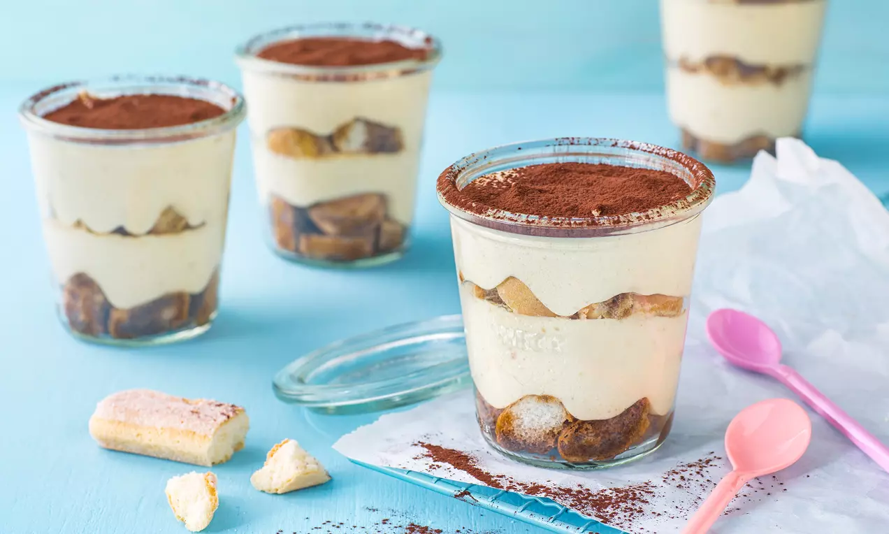 Schichtdessert aus hellgelber Creme und braunen Löffelbiskuits in einem klaren Glas mit Kakao bestäubt, daneben bunte Löffel und Krümel