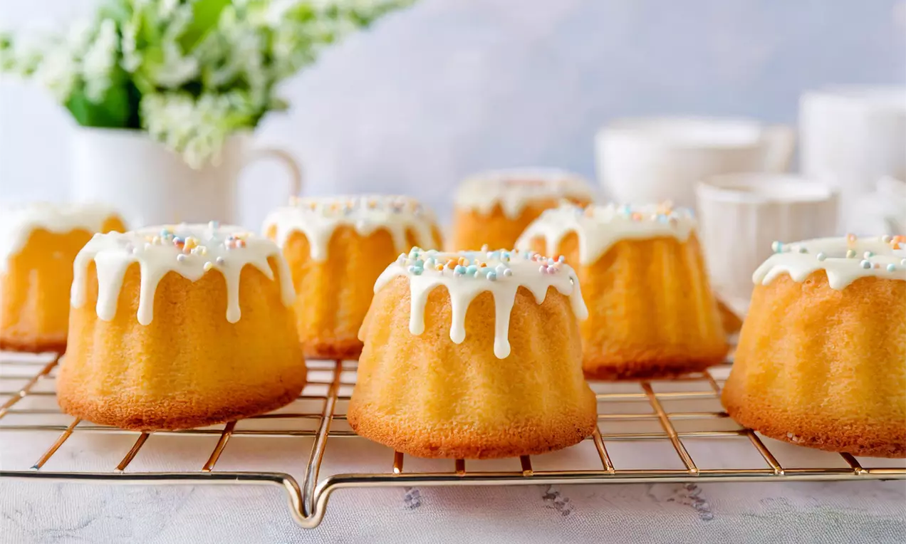 Mini Gugelhupf dorati con una glassa bianca che cola delicatamente e granelli colorati, disposti su una griglia dorata