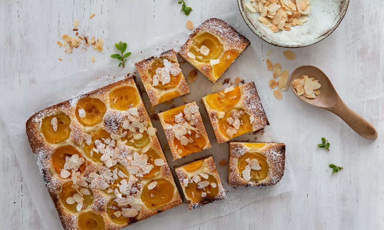 Gâteau moelleux rectangulaire doré garni de demi-abricots juteux, d'amandes effilées et de sucre glace étalé sur une table en bois clair