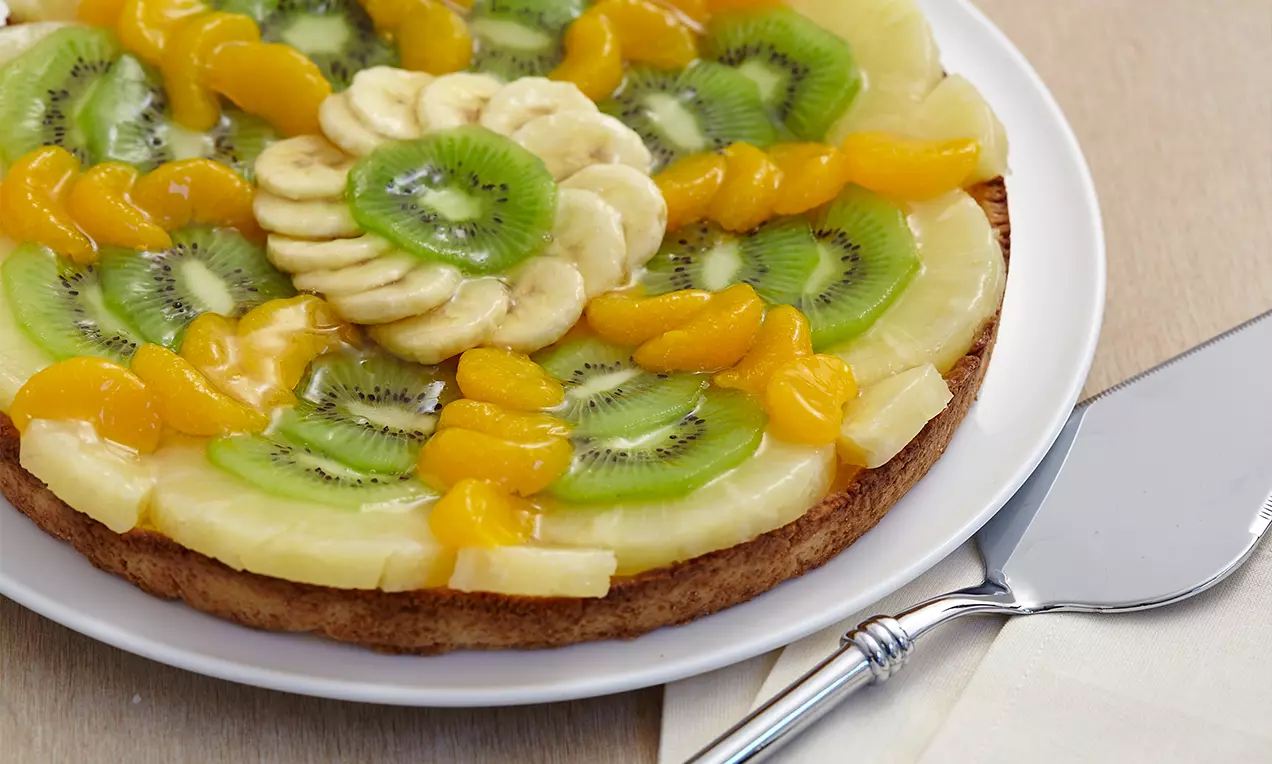 Torta di frutta su base dorata croccante, con fette di kiwi verde brillante, mandarini arancioni succosi, ananas chiaro e banana disposti a corona