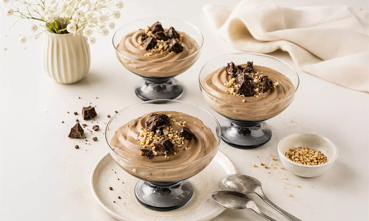 Luftig-cremes Mousse mit zarter Schokoladenfarbe in Gläsern, garniert mit Schokoladenstückchen und gehackten Nüssen, auf einem hellen Tisch., AI-labeled image