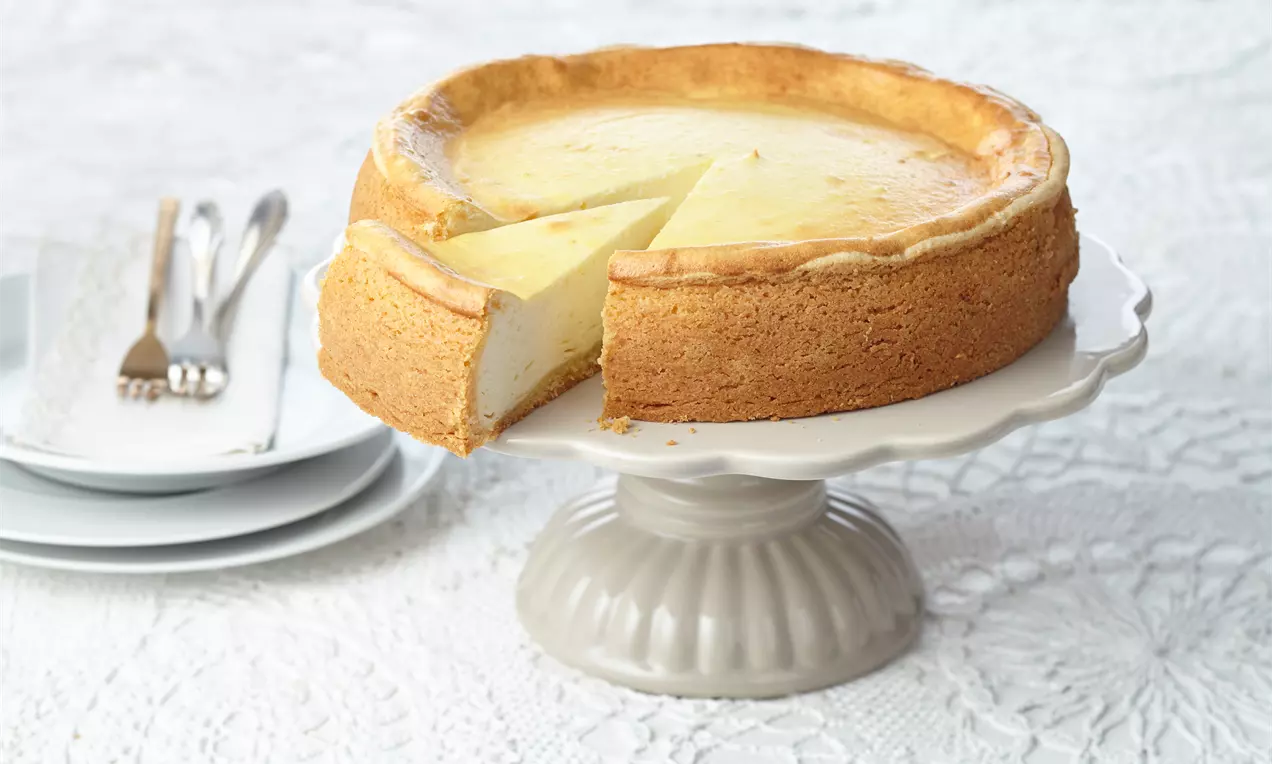 Klassischer deutscher Käsekuchen mit goldbraunem Rand und leicht cremiger Füllung, serviert auf weißer Tortenplatte mit Besteckset