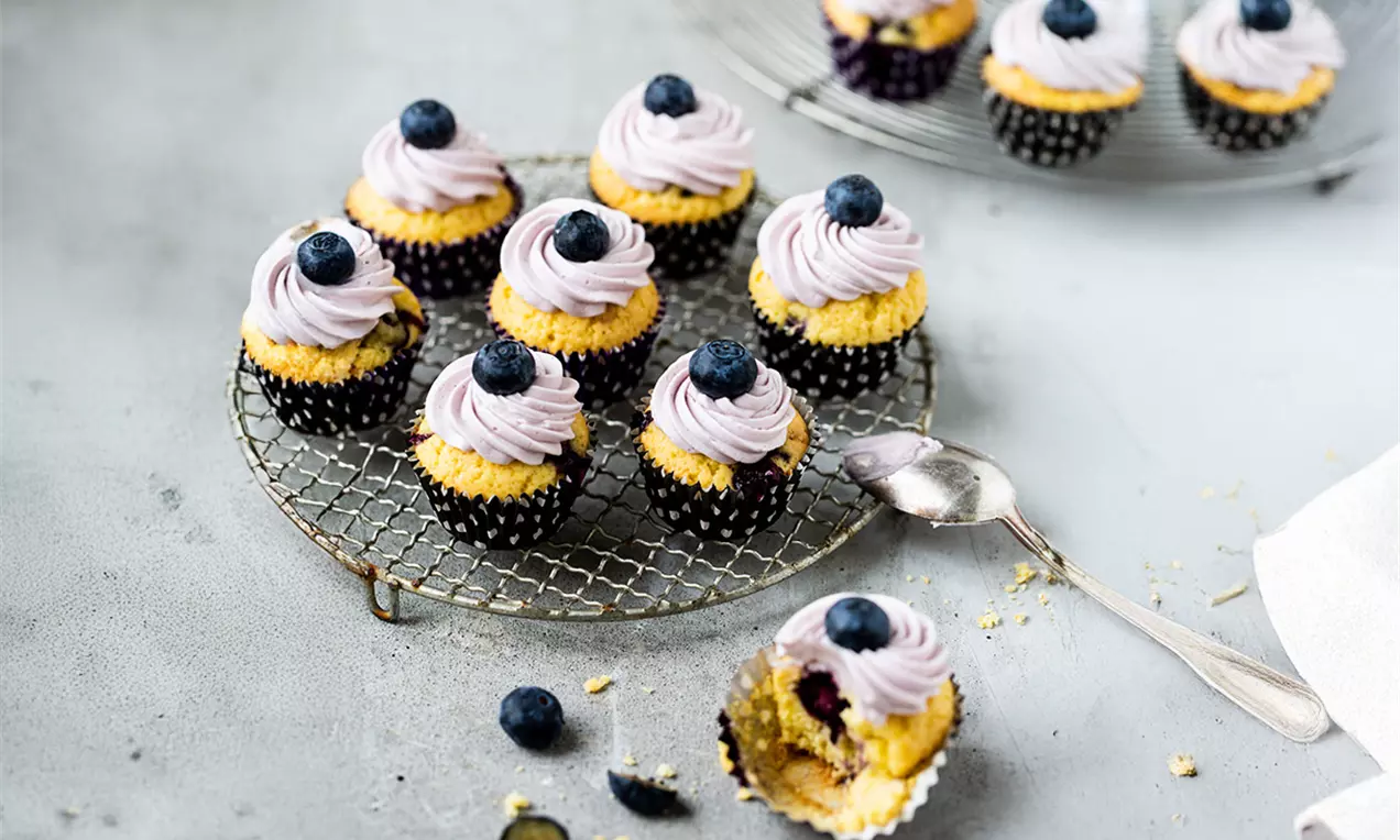 Kleine, goldgelbe Mini-Muffins mit lila Cremehäubchen und einzelnen Blaubeeren, arrangiert auf einem metallenen Kuchengitter, AI-labeled image
