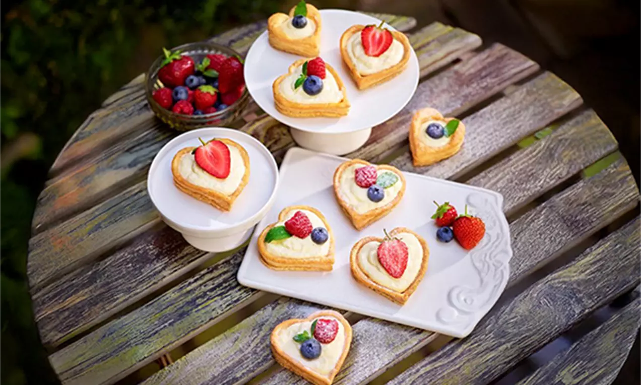 Herzförmige Blätterteiggebäcke mit goldbrauner Kruste, belegt mit frischen Beeren und grünen Blättern, arrangiert auf Tellern und Holz