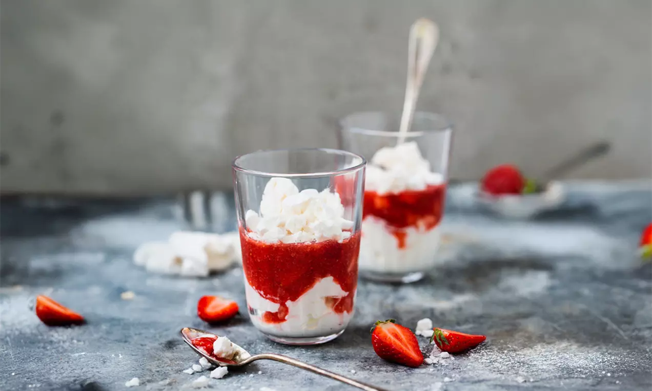 Erdbeerdessert mit leuchtend roter Fruchtschicht, heller Creme und knusprigem Baiser in gläsernen Bechern auf dunklem Tisch mit frischen Erdbeeren, AI-labeled image
