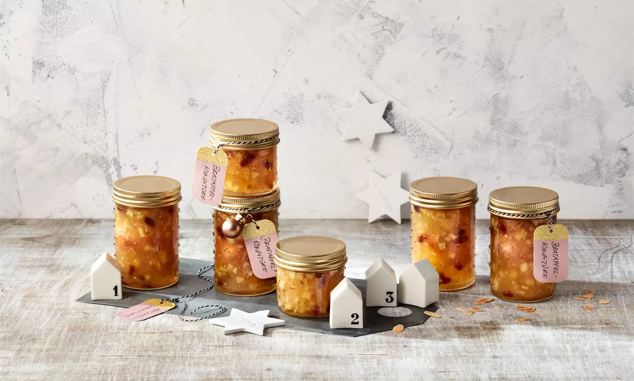 Bratapfel-Konfitüre in golden verschraubten Gläsern mit sichtbaren Fruchtstückchen, auf Holzplatte dekorativ arrangiert mit Sternen und Zahlenhäusern