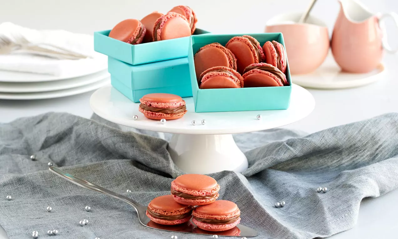 Runde, rotbraune Macarons mit glatter Oberfläche und weißlichen Nuss-Nougat-Rändern, drapiert auf einer weißen Tortenplatte mit hellblauen Schachteln