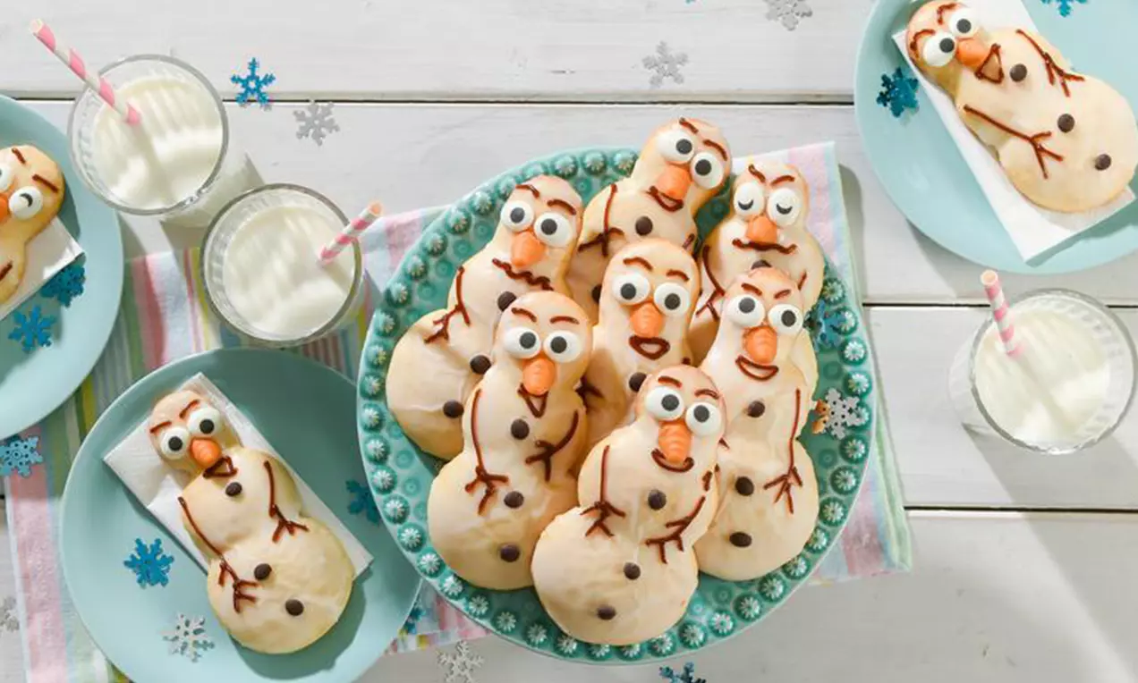 Bonshommes de neige en pâte dorée aux détails en chocolat et glaçage blanc, disposés sur une assiette turquoise entourée de verres de lait
