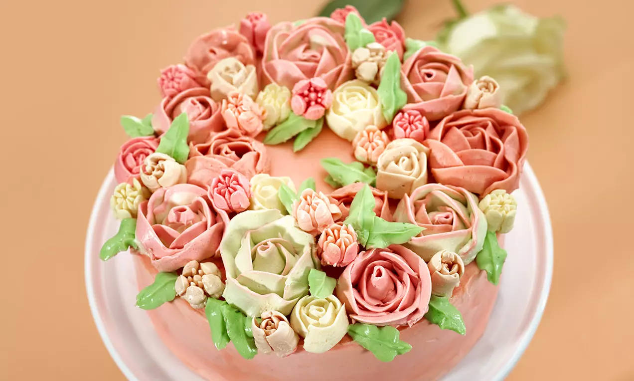Runde Torte mit rosa Buttercreme, verziert mit filigranen Blüten aus Creme in Rosétönen und Grün, arrangiert auf einer weißen Tortenplatte