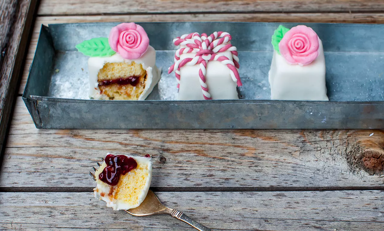 Kleine Würfel mit weißem Zuckerguss, dekoriert mit rosafarbenen Marzipanrosen und grünen Blättern, in einer metallenen Schale
