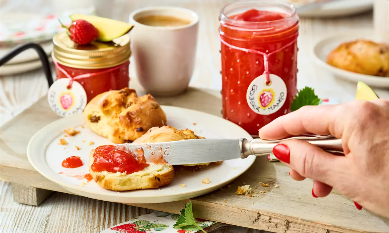 Erdbeer-Mango-Marmelade in einem offenen Glas, daneben goldbraune Brötchen auf einem Teller, bestrichen mit der leuchtend roten Marmelade