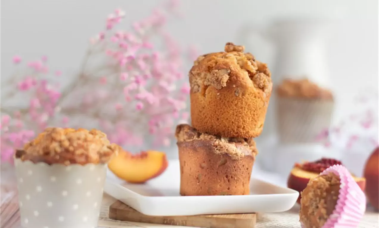 Goldbraune Muffins mit knusprigen Streuseln, locker gestapelt auf weißem Teller, umgeben von rosa Blumen und frischen Pfirsichstücken