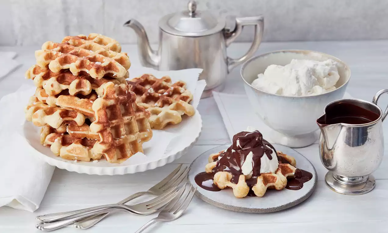 Belgische Waffeln golden und luftig auf weißem Teller gestapelt, daneben Waffel mit Sahnehäubchen und Schokoladensoße auf grauem Teller