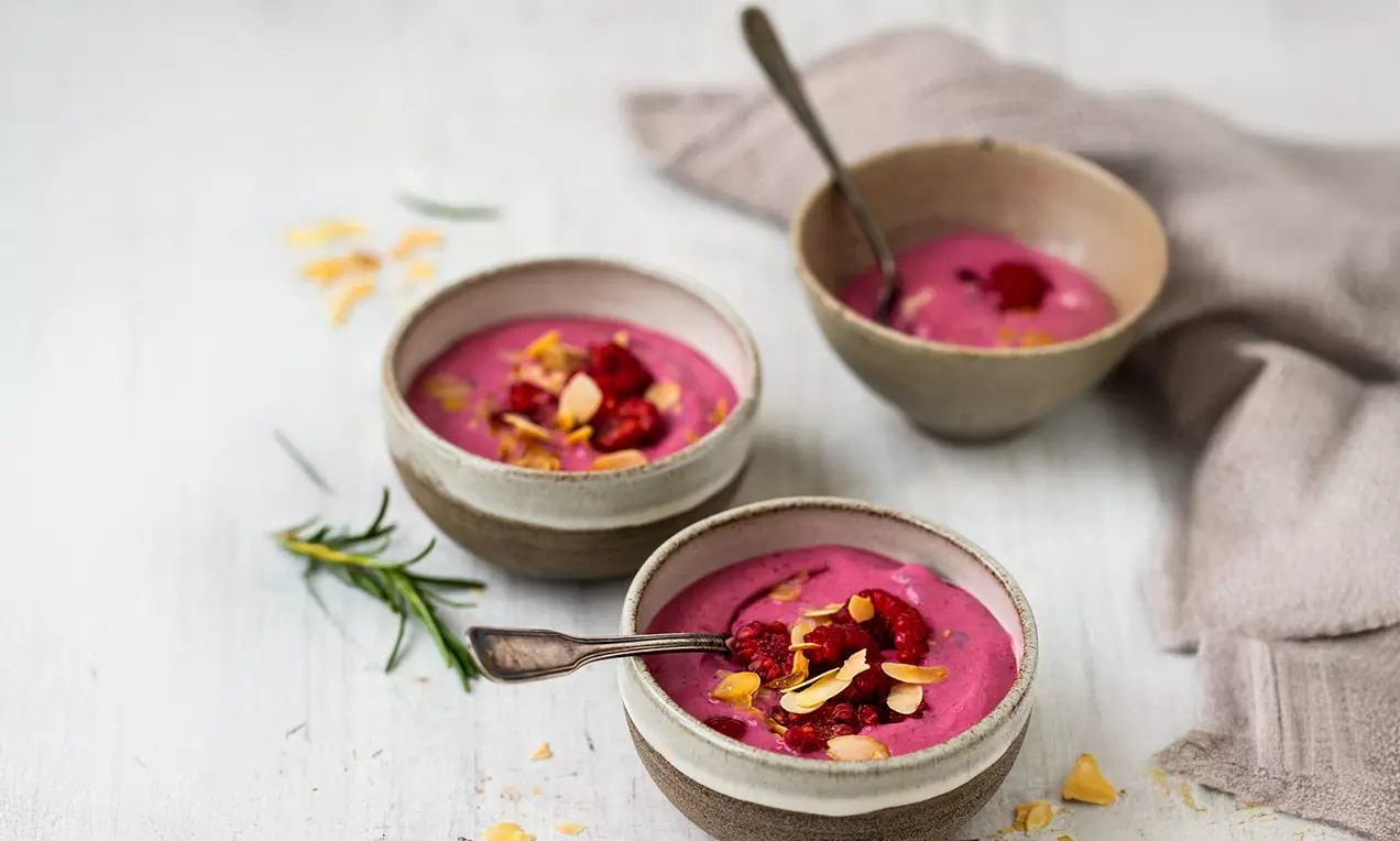 Leuchtend pinke Himbeercreme in beigefarbenen Schalen, garniert mit frischen Himbeeren und Mandelsplittern, auf hellem Holztisch drapiert