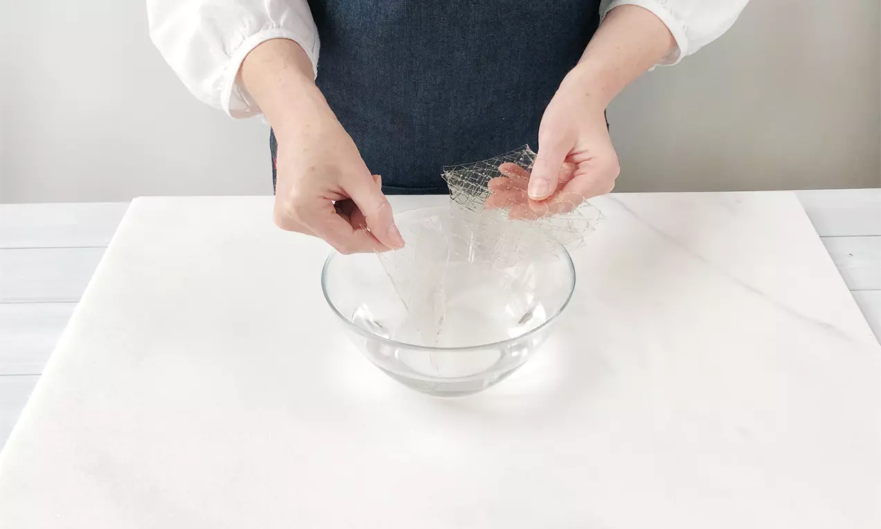Eine Person in weißer Bluse und dunkler Schürze weicht klare Gelatineblätter in einer geschwungenen Glasschüssel auf weißer Arbeitsfläche ein