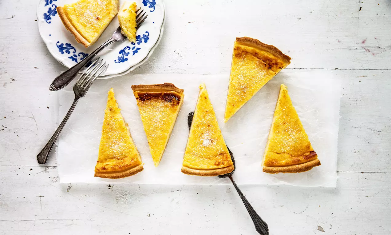 Fette triangolari di torta alla crema dorata con crosta leggermente scura distribuite su carta bianca, una fetta su piattino decorato con posate
