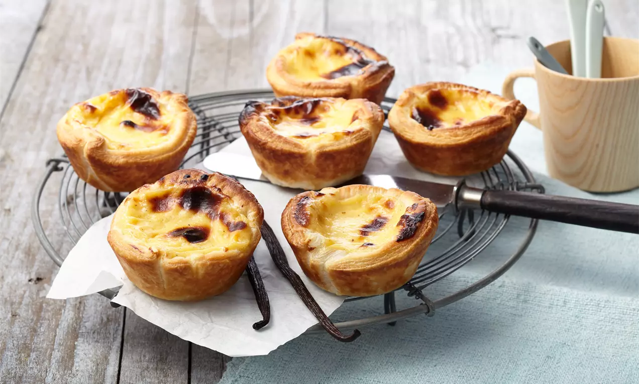 Goldbraun gebackene Pasteis de Nata mit karamellisierter Oberfläche auf einem runden Gitterrost, begleitet von Vanilleschoten und Papier