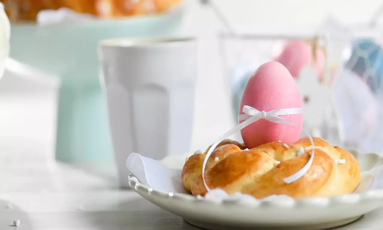 Petites couronnes dorées en pâte moelleuse ornées d’un œuf rose avec un ruban blanc, posées sur une assiette beige délicatement dentelée