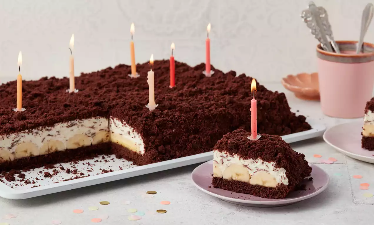 Maulwurfkuchen vom Blech mit dunkler Krümelschicht, heller Bananen-Sahne-Füllung und bunten Kerzen auf Tellern und Tablett angerichtet