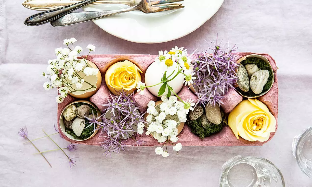 Cartone rosa con fiori bianchi, gialli e viola, gusci d’uovo e pietre su muschio verde, disposto su una tovaglia lilla con piatti e posate