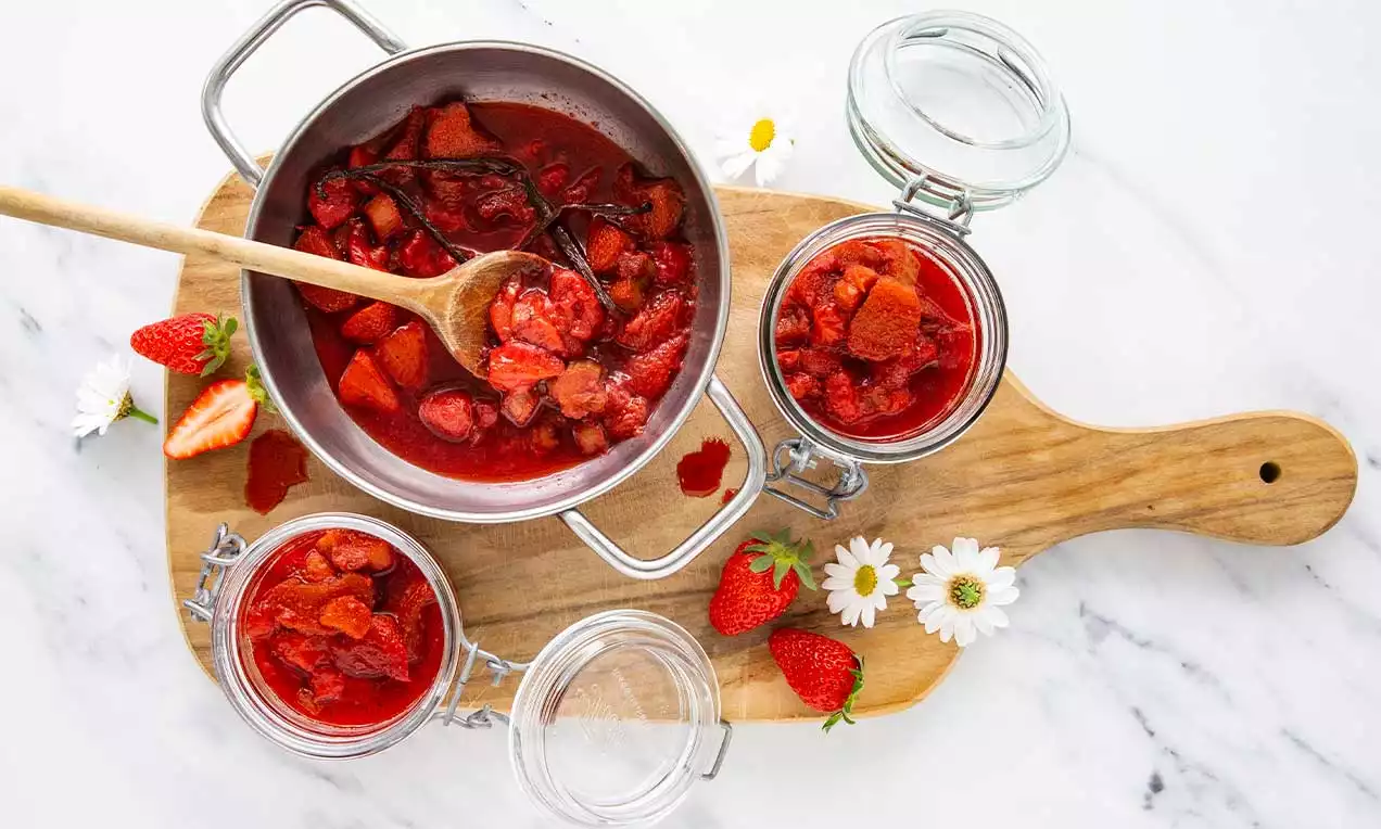 Erdbeer-Rhabarber-Kompott tiefrot und fruchtig, mit Vanillestangen in einem Topf und drei Gläsern auf einem Holzbrett mit frischen Erdbeeren und Gänseblümchen