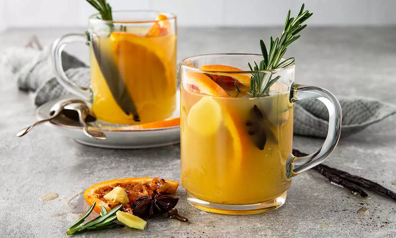 Hellgelber Orangenglühwein in klaren Glastassen mit Rosmarinzweig, Orangenscheiben und Gewürzen auf grauem Tisch mit dekorativer Beilage