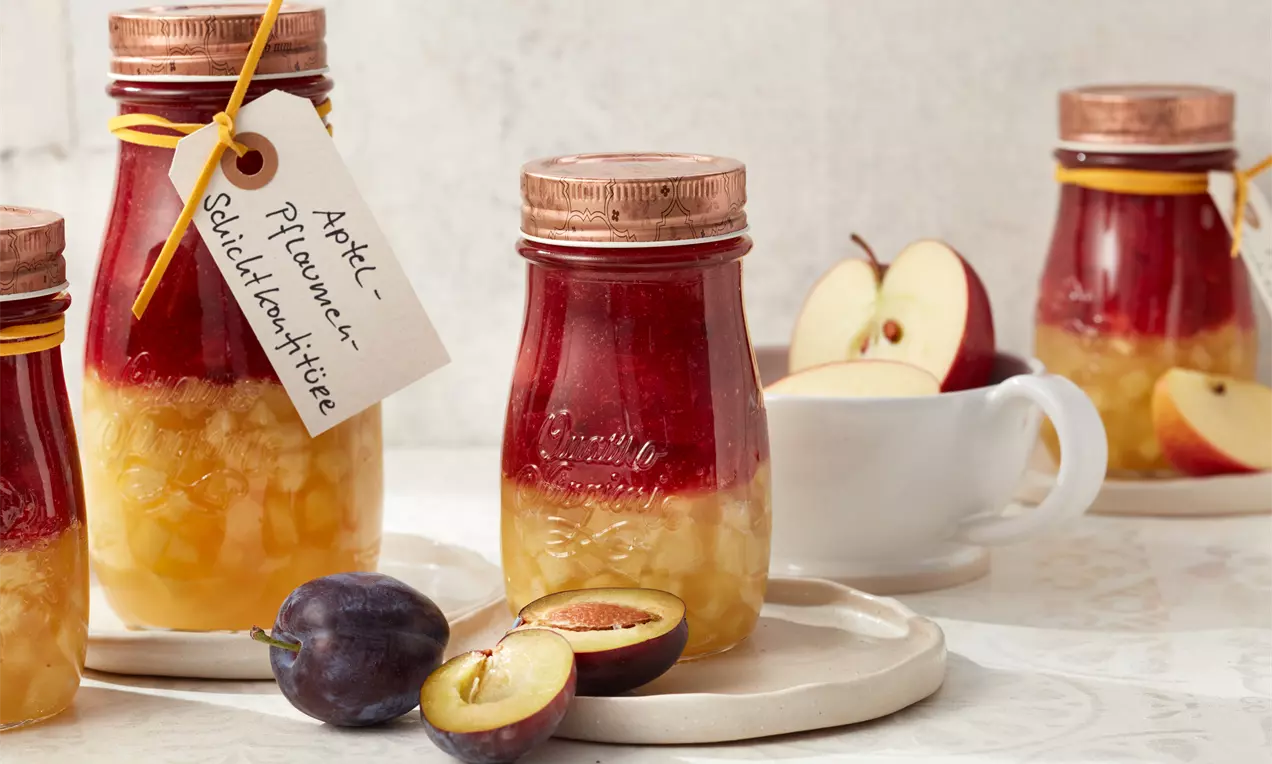 Confiture en couches rouge foncé et dorée dans un pot de verre avec couvercle cuivré, étiquetée à la ficelle jaune, prune et pomme à côté