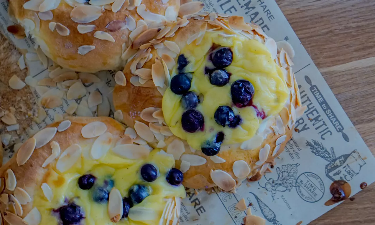 Goldbraune Hefebrötchen mit Vanillecreme, frischen Blaubeeren und gerösteten Mandelblättchen auf bedrucktem Papier angerichtet