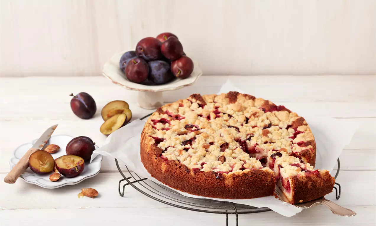 Gâteau rond aux prunes violet foncé et jaunes sous une couche de crumble doré sur une grille entouré de fruits frais et coupés