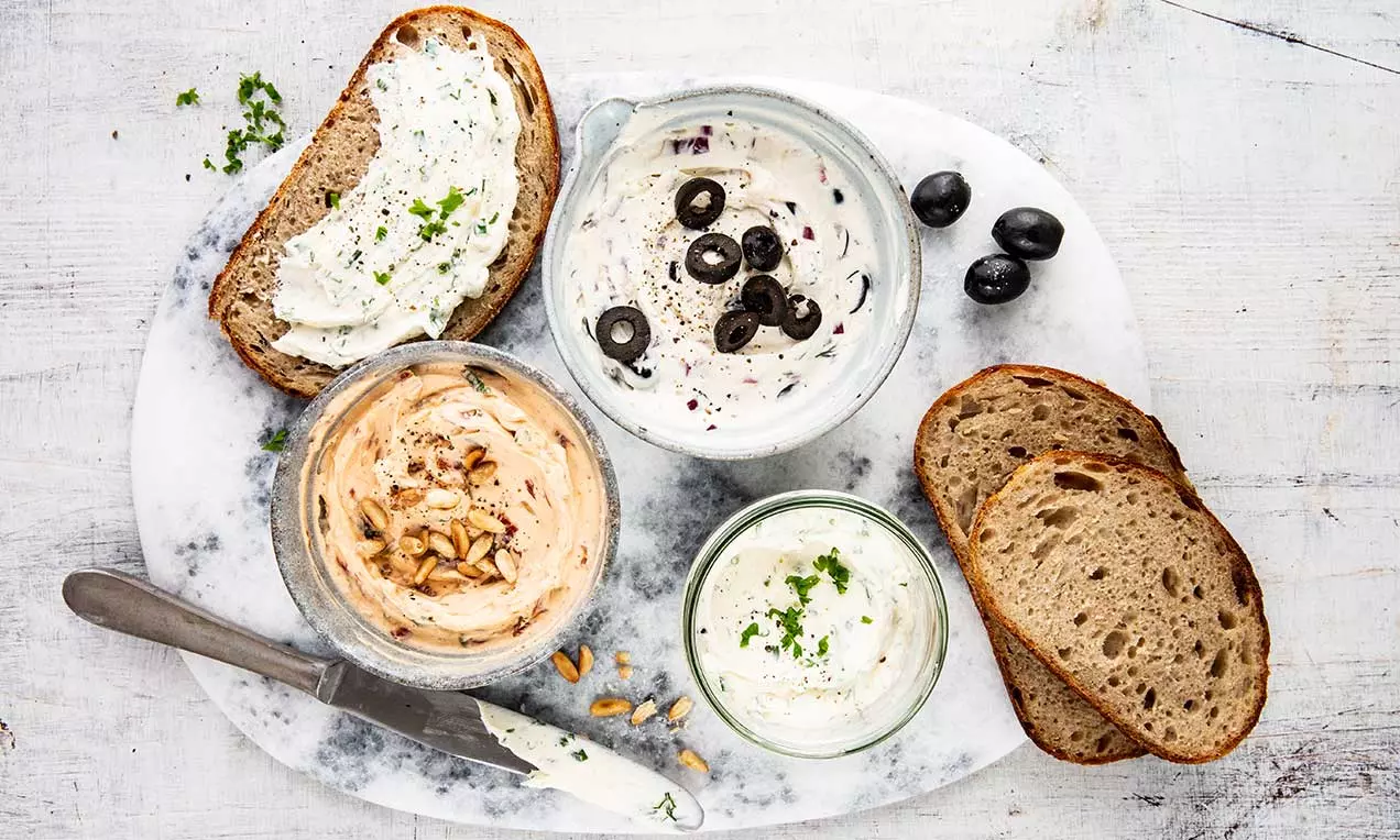 Tris di creme spalmabili in ciotole di vetro, con erbe verdi, olive nere e pinoli dorati, accanto a fette di pane rustico su piatto marmoreo