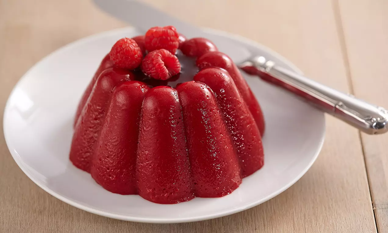 Dessert rosso brillante a forma di cupola ondulata decorato con lamponi freschi su un piatto bianco, con un coltello argentato accanto