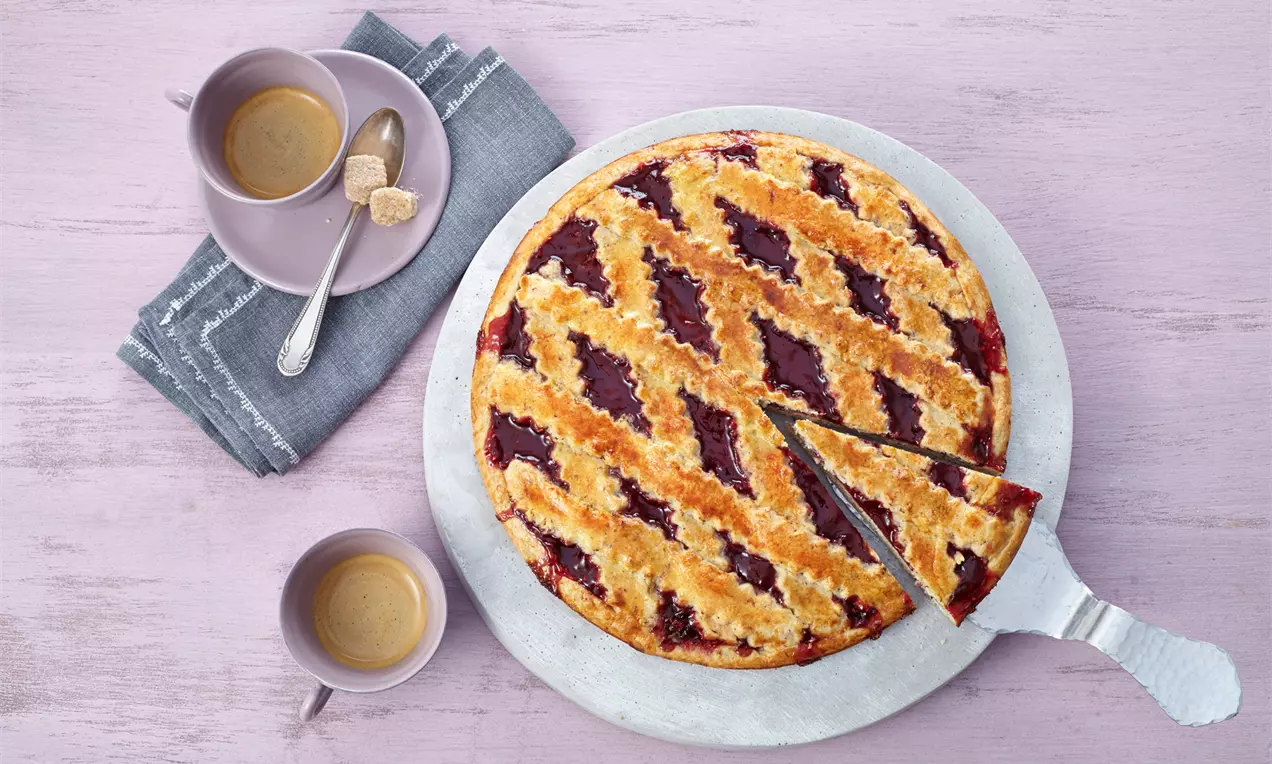 Runde Linzer Torte mit goldbrauner Teigoberfläche und rotem Marmeladenraster auf weißem Teller, daneben Tassen mit Kaffee und Zuckerwürfeln