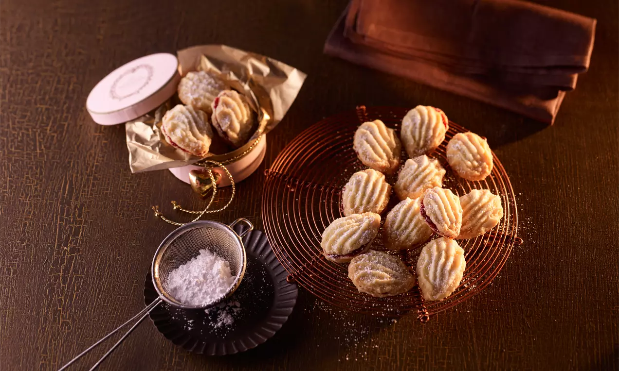 Biscotti dorati con forma ondulata e crema al centro, disposti su una griglia rossa accanto a una scatola decorata e un setaccio con zucchero
