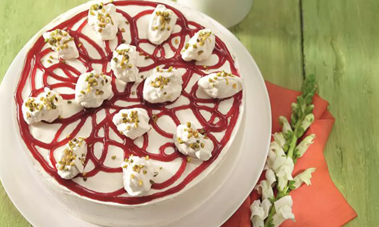 Gâteau rond garni de chantilly blanche en touffes, filaments rouges brillants et éclats de pistache sur une assiette blanche