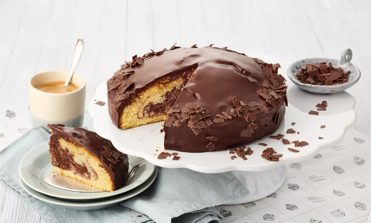 Runder Marmorkuchen mit glänzender Schokoladenglasur, dekoriert mit Schokoraspeln, ein angeschnittenes Stück auf Teller mit Gabel daneben