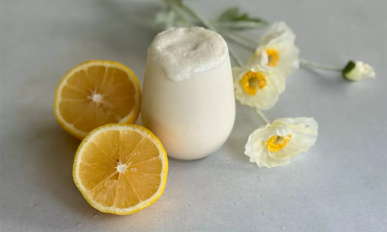 Ein cremiges, hellgelbes Getränk mit schaumiger Spitze in einem glatten Glas, umgeben von halben Zitronen und zarten weißen Blüten