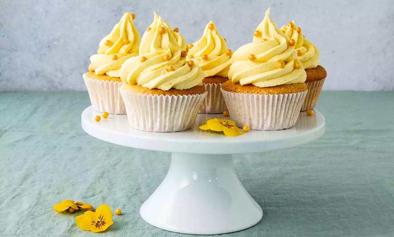 Vier Mango-Vanille-Cupcakes mit goldgelber Cremehaube und karamellfarbener Deko auf weißer Tortenplatte, umrahmt von gelben Blüten