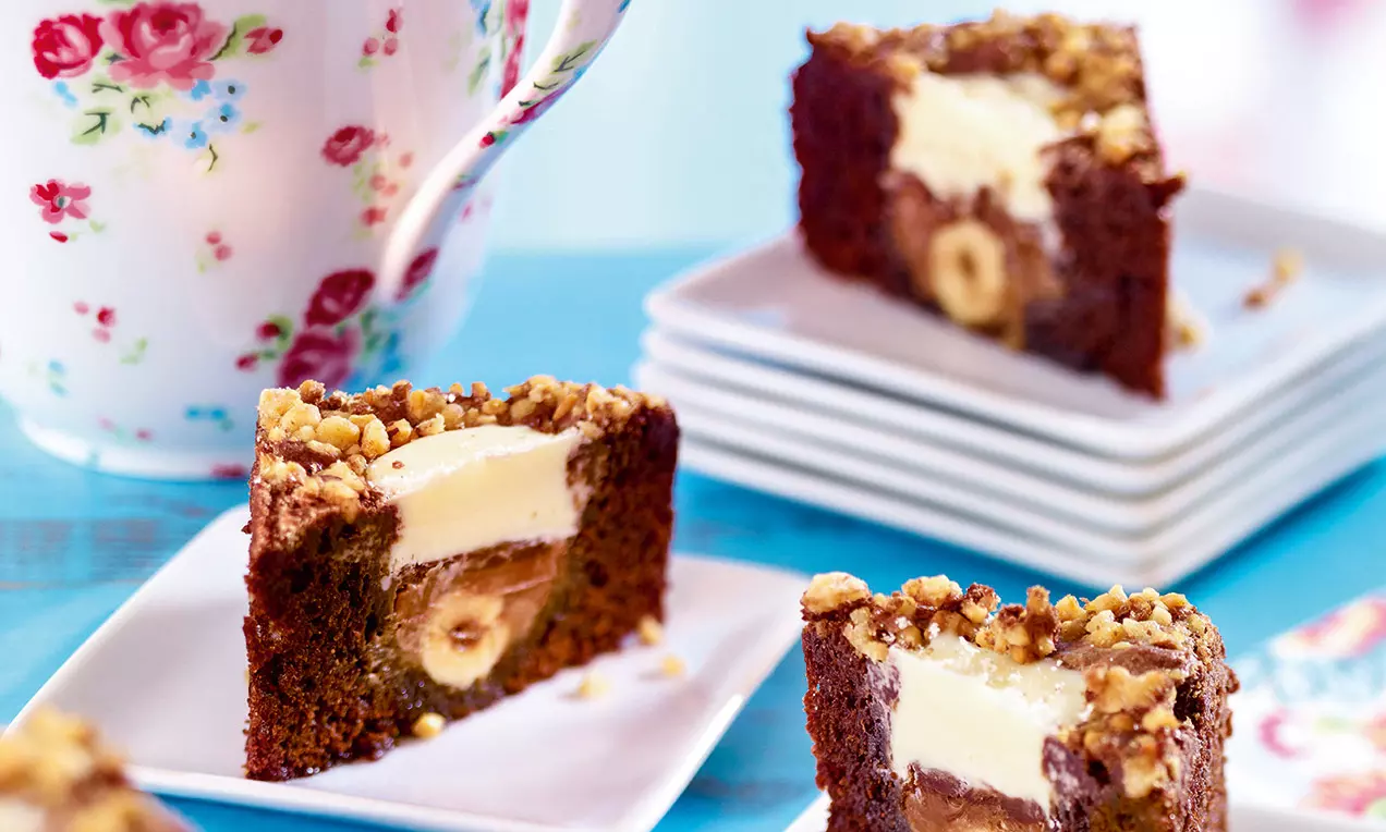 Quadrotti di brownie scuri con cuore di crema chiara e nocciole intere, guarniti di granella dorata su piattini bianchi accanto a una tazza floreale