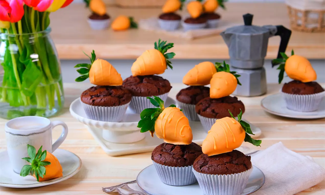Muffin al cioccolato scuro con superficie morbida, decorati con fragole immerse in cioccolato arancione e foglie verdi su piatti e tavolo di legno
