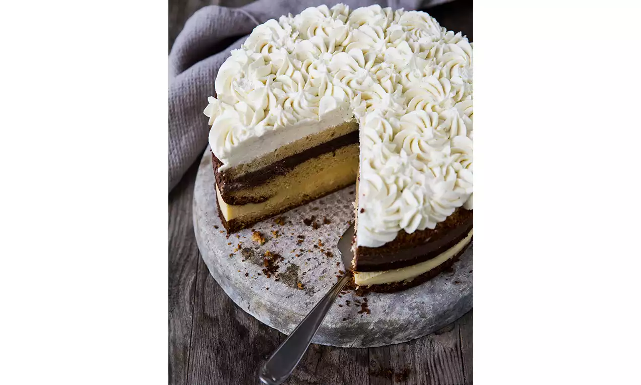 Gâteau en couches avec crème mascarpone ivoire en volutes sur le dessus, base brune et biscuit doré, présenté sur une planche ronde