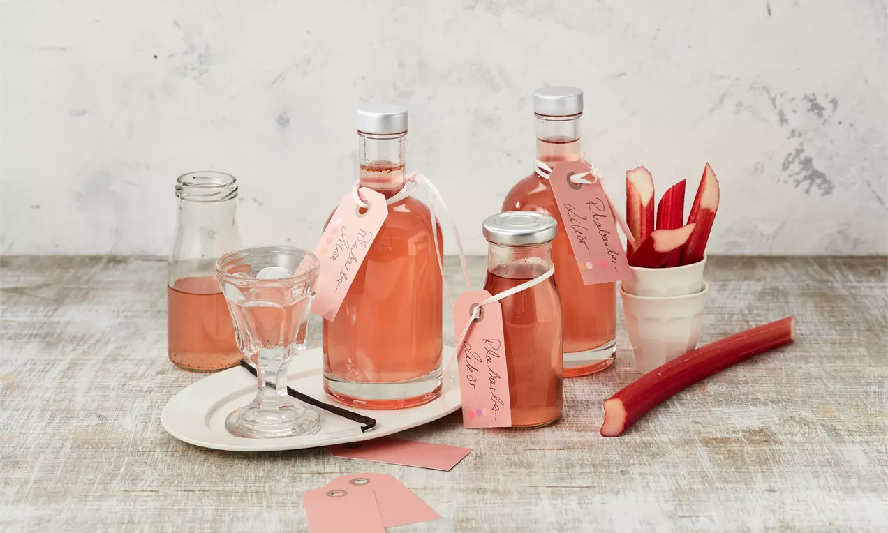 Mehrere Glasflaschen mit rosafarbenem Rhabarber-Likör, dekoriert mit rosigen Etiketten, arrangiert neben einem Glas und frischem Rhabarber