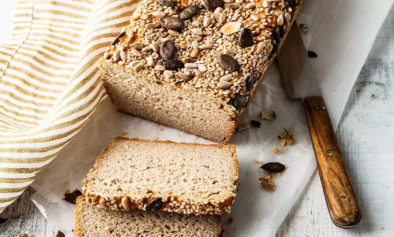 Pane gluten-free dalla crosta dorata cosparsa di semi misti, con fette morbide e compatte su carta bianca accanto a un coltello di legno