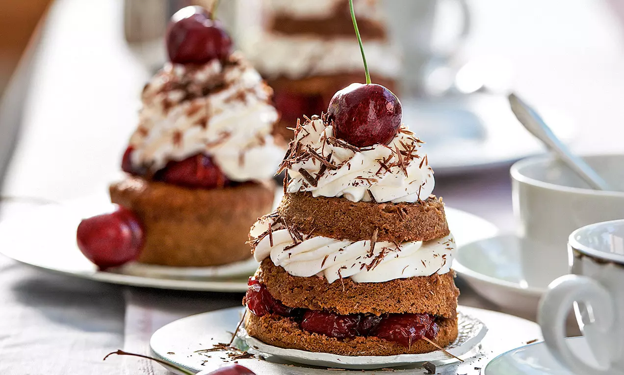 Tortino a strati con base scura, crema bianca soffice, riccioli di cioccolato e ciliegia rossa con picciolo su un piatto chiaro