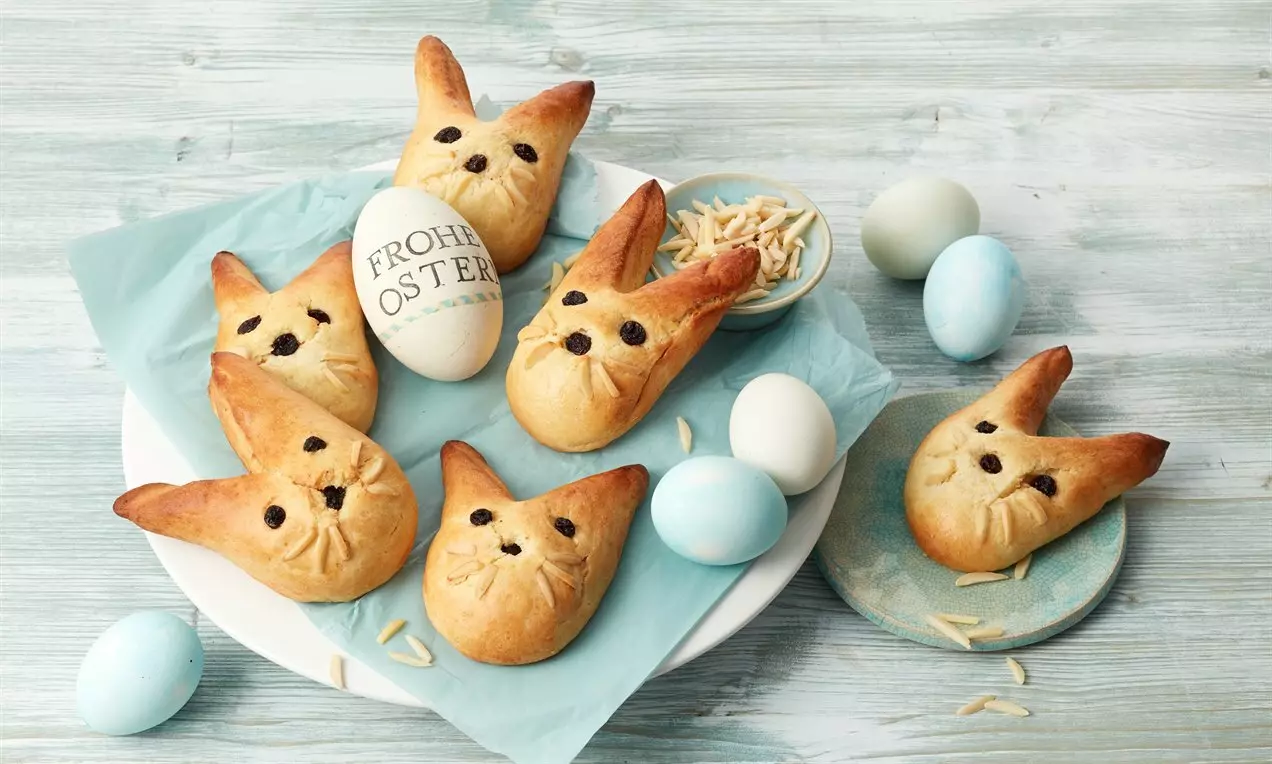 Goldbraune, hasenförmige Gebäckstücke mit Rosinenaugen auf hellblauem Papier, umgeben von pastellfarbenen Ostereiern und Mandelsplittern