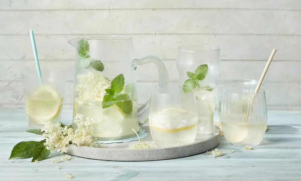 Kühle Holunderblüten-Limonade in Gläsern und einer Karaffe, mit Zitronenscheiben, Minzblättern und Holunderblüten auf hellem Tablett arrangiert