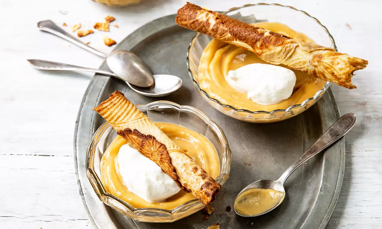 Crema caramello lucida in coppette di vetro con panna bianca soffice e cialde dorate sottili croccanti su vassoio con cucchiaini argento